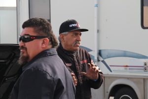 An older Hispanic gentleman in a black racing suit talking