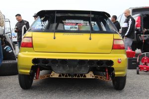 Rear of a drag racing prepared Honda Civic hatchback, mustard yellow color, showing bumper cutouts, subframe brace, custom rear wing, aftermarket shocks