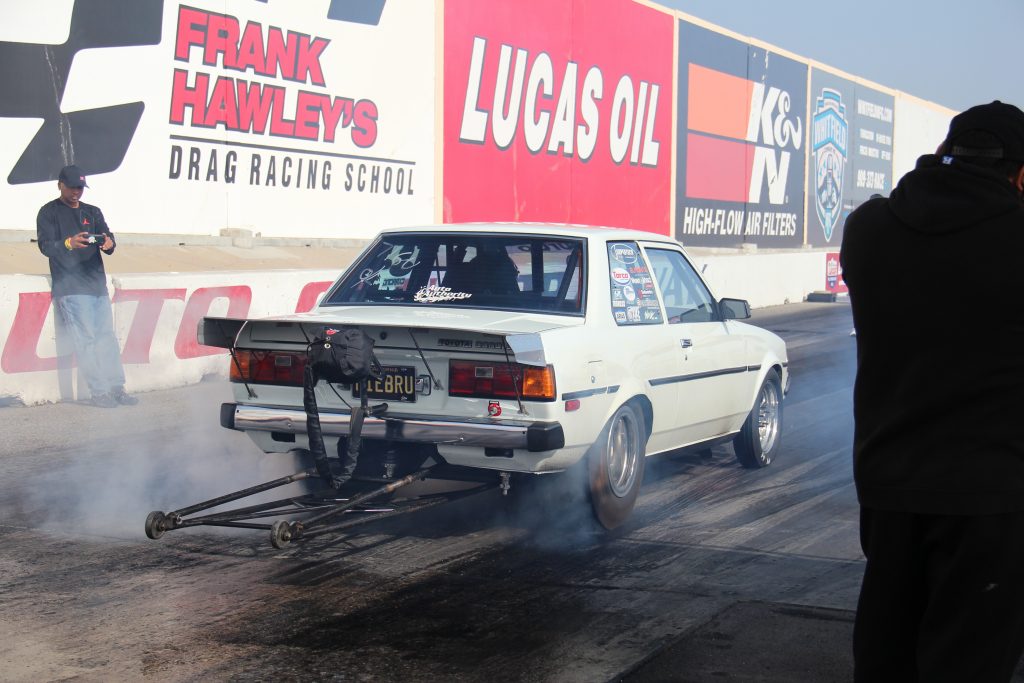 An older Toyota Corolla, with parachute and wheeli bars, does a burnout on a drag strip