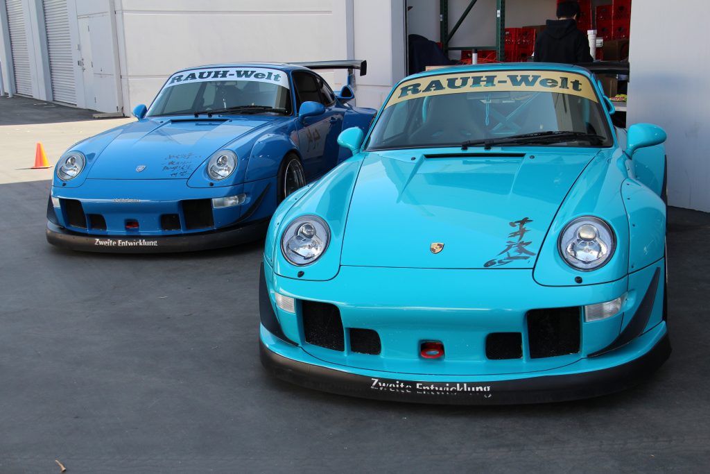 Two RWB Porsches parked outside a warehouse: Meguri-Ai, a blue Porsche 993 and Shingen, a teal Porsche 993.