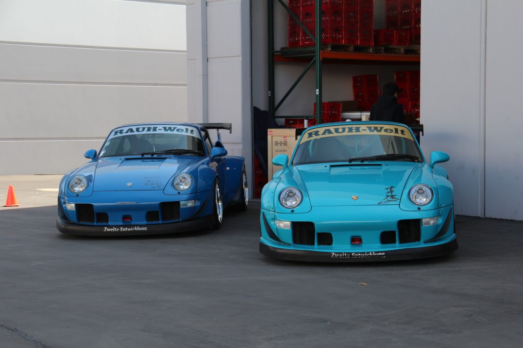Two RWB Porsches parked outside a warehouse: Meguri-Ai, a blue Porsche 993 and Shingen, a teal Porsche 993.